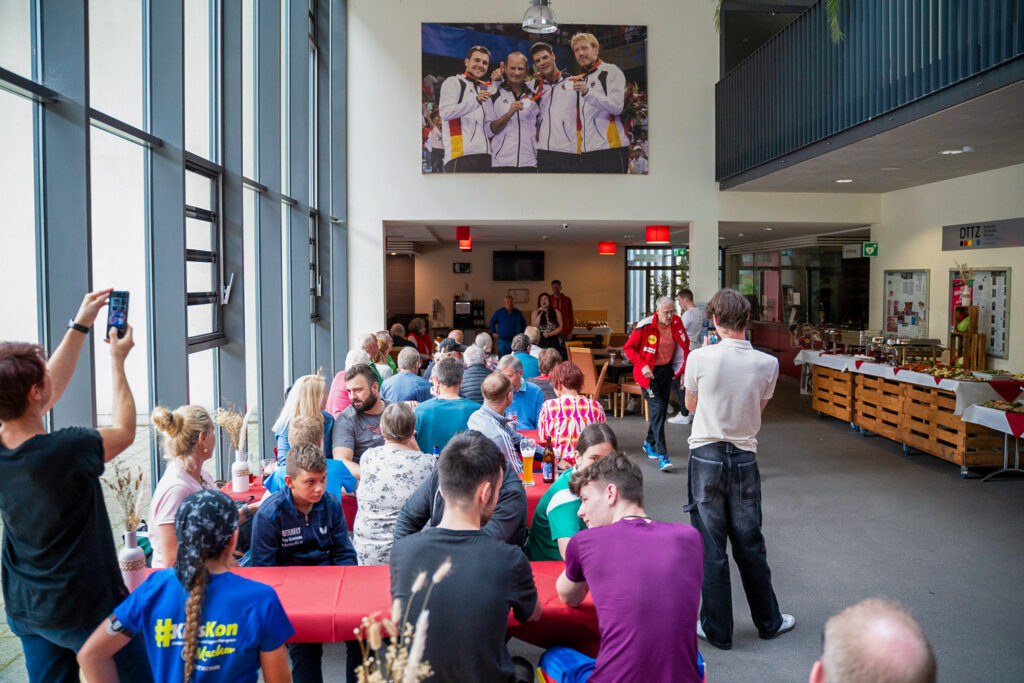 Menschen in einer Halle sitzen und sprechen, große Fotografie im Hintergrund