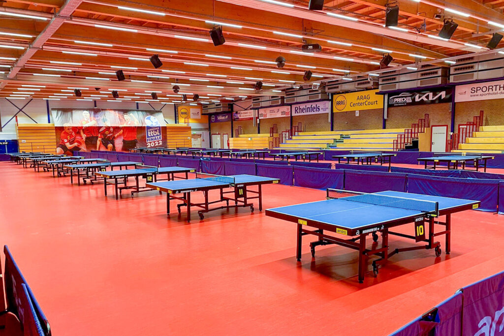 Leere Tischtennishalle mit mehreren Tischen und gelben Tribünen