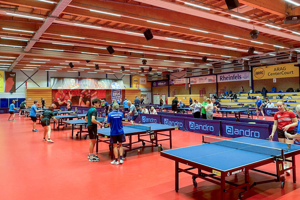 Tischtennisturnier in großer Sporthalle mit Spielern an mehreren Tischen